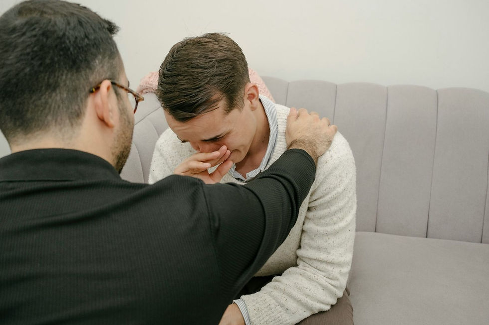 A man in a sweater sits on a couch, crying. Another person beside him in black comforts him by placing a hand on his shoulder.