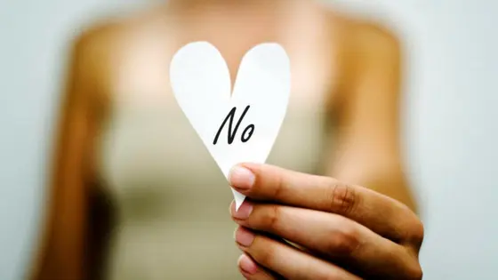 A person holds a white heart-shaped paper with "No" written on it. The background is blurred, emphasizing the paper and hand.
