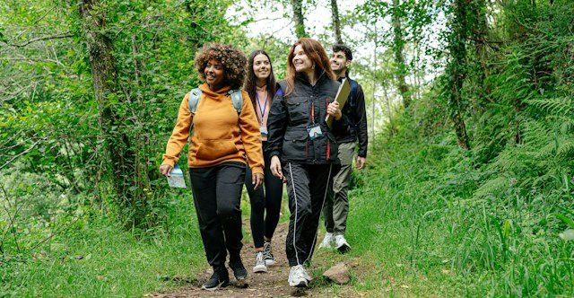 Four friends hike a green forest trail, smiling and chatting, with backpacks and casual outdoor clothing.