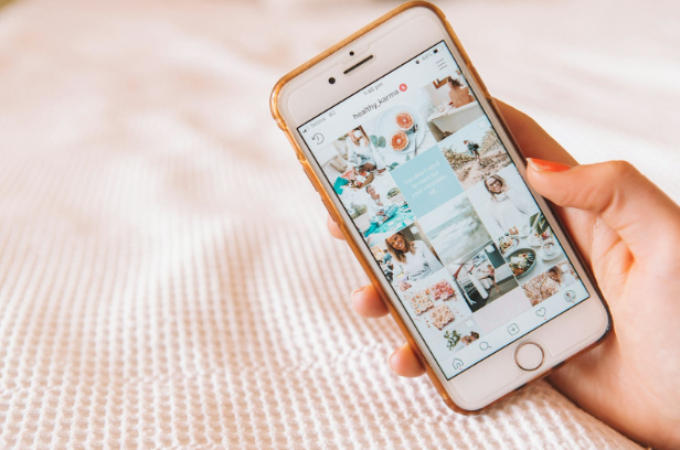 A hand holds a smartphone displaying an Instagram profile with photos. The background is a soft, white textured surface.
