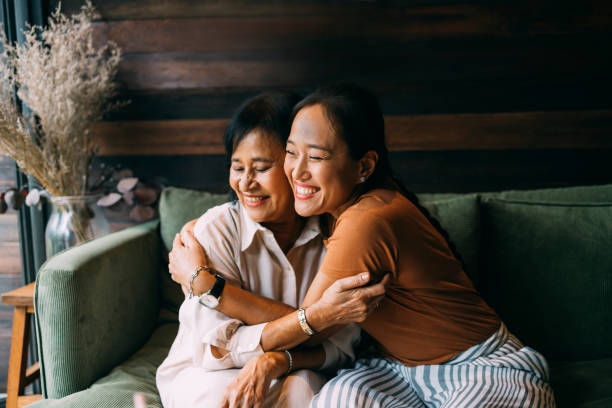 Two women hugging and smiling on a green couch, in a cozy setting. One wears a white shirt, the other a brown top. Warm, joyful mood.