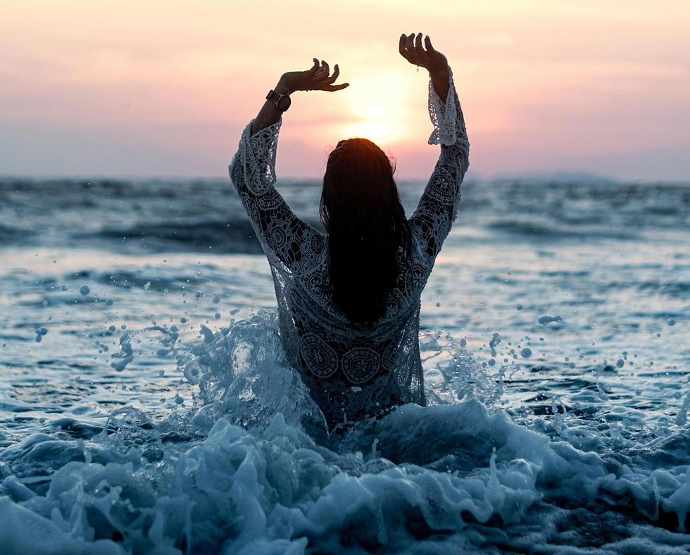 Woman in lace garment raises arms in ocean waves at sunset. Water splashes around, creating a serene, joyful atmosphere.