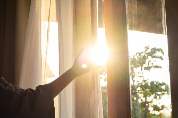 Hand opening a curtain with sunlight streaming in through a window. Trees visible outside, creating a calm, serene morning ambiance.