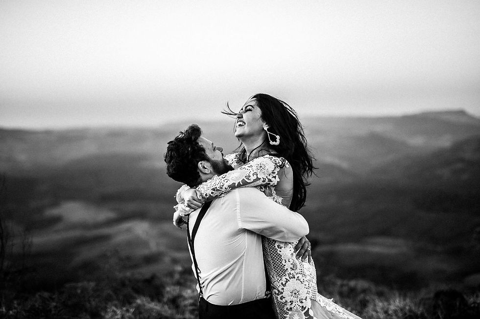 a joyous couple embracing outdoors, with the woman laughing and the man lifting her, set against a scenic mountainous backdrop in black and white.