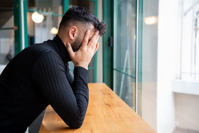 Man in a black sweater sits at a wooden table, covering his face with his hands, looking distressed. Glass door in the background.