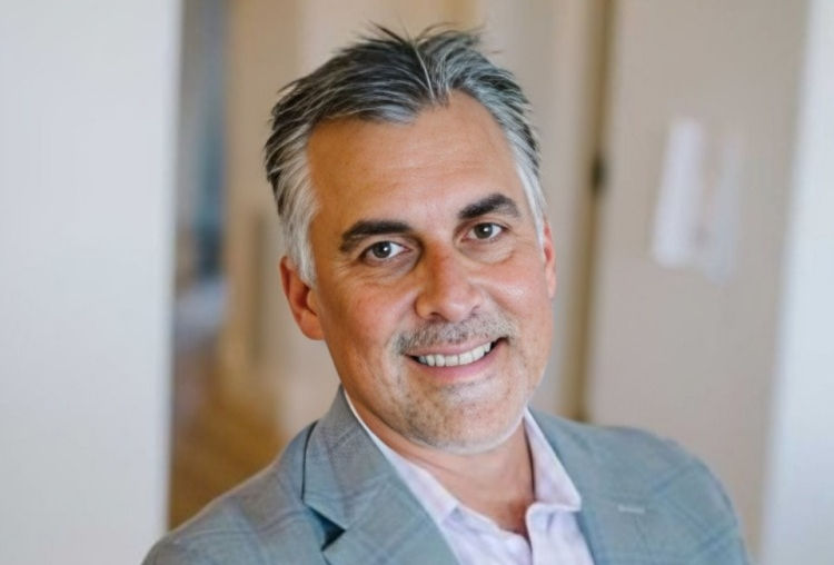 Smiling man in a gray blazer stands indoors against a blurred background, exuding a friendly, approachable mood.