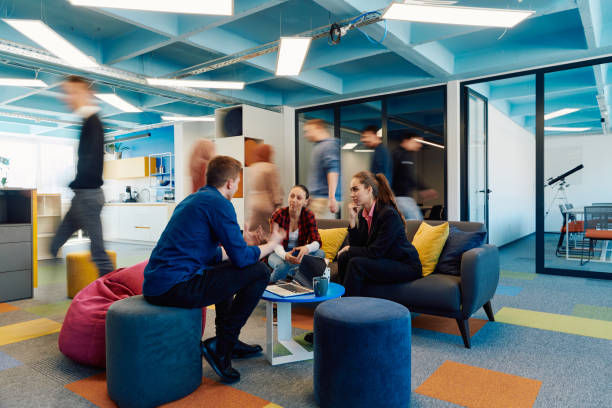 People in a brightly colored office lounge, engaged in discussion on sofas, while others walk past. Blue ceiling, colorful carpet, relaxed mood.