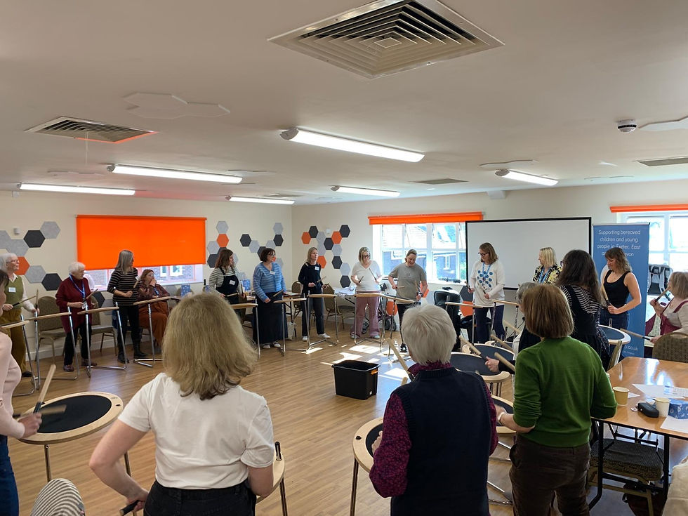 People in a bright classroom, standing in a circle, holding drumsticks. Orange window shades, hexagonal wall designs, and a banner visible.