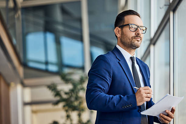 Man in a blue suit and glasses holds papers and a pen in a modern office, looking thoughtful.