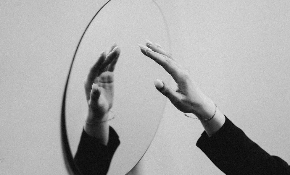 Black-and-white hands reaching toward an oval mirror, with a reflected hand and a moody, minimalist background.
