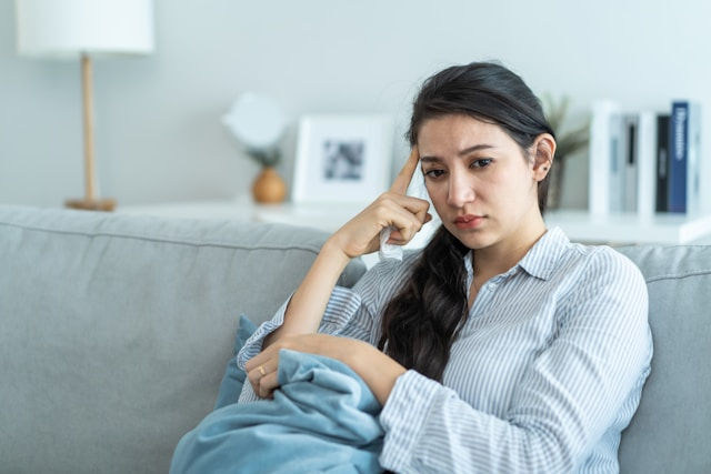 A woman sits on a couch, looking deep in thought with a concerned expression while holding a blanket.