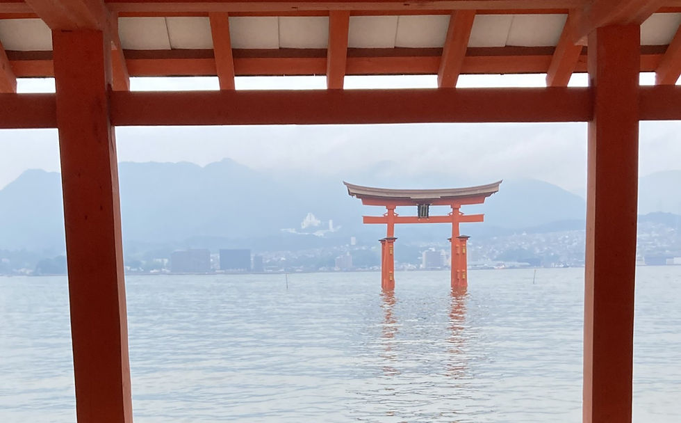 Red torii gate standing in calm water, framed by shrine beams, with misty mountains and a hazy lakeside town beyond.