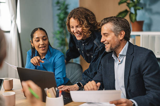 Three coworkers smile and collaborate around a laptop in a bright office, with notes and mugs on the table.
