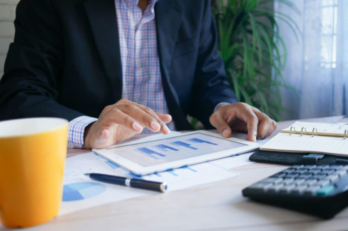 Person in a suit uses a tablet displaying graphs at a desk with documents, a yellow mug, pen, and calculator. Indoor office setting.