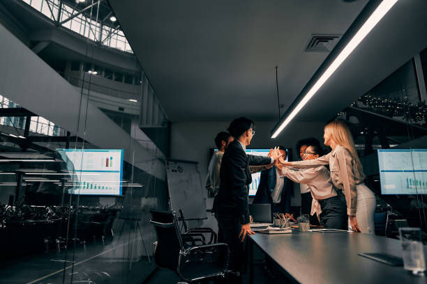 Business team in a modern office celebrating. Five people high-fiving, surrounded by screens displaying graphs. Energetic and collaborative mood.