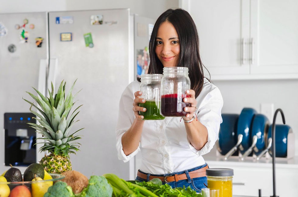 Smiling woman in a bright kitchen holds two jars of green and red juice beside fresh fruits and vegetables on the counter