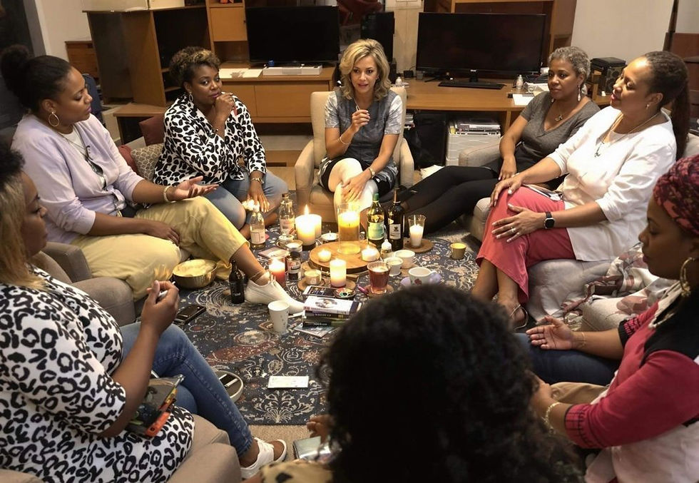Eight women gather in a cozy room, sitting in a circle with drinks and candles on a patterned rug, engaged in conversation.