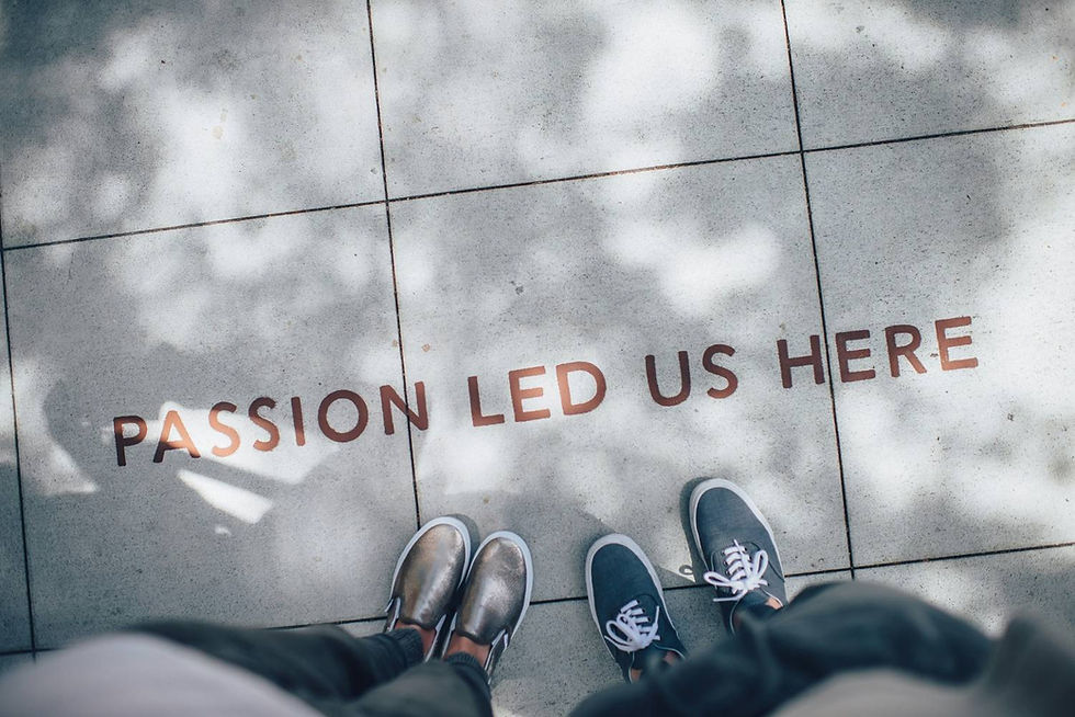 Two pairs of shoes on a sunlit sidewalk with "PASSION LED US HERE" written in bold letters. Shadows create a serene mood.