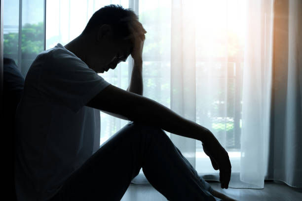 Silhouette of a man sitting with head in hand, appearing distressed. Sunlight pours through sheer curtains in a dimly lit room.
