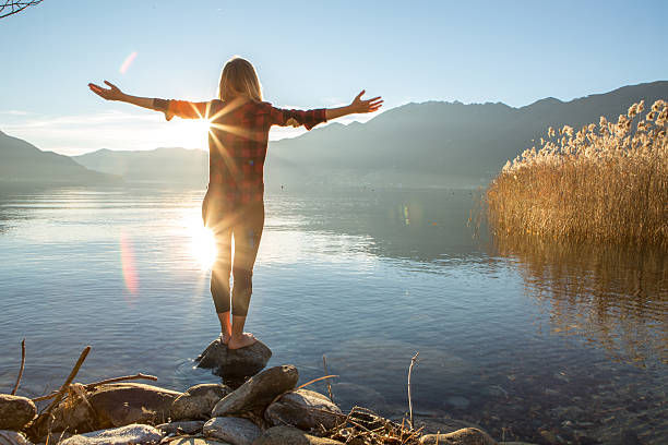Woman in plaid shirt stands on rocks by a calm lake at sunrise, arms outstretched, with mountains and reeds in the background.
