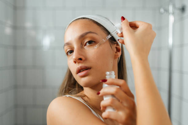 Woman applying serum under her eye with a dropper in a tiled bathroom. She wears a white headband, expressing focus and self-care.