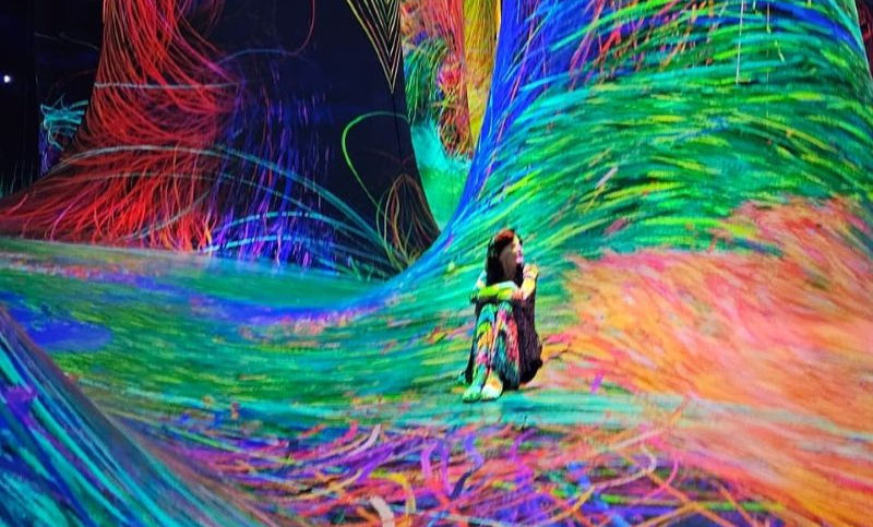 A woman sits cross-legged in a vivid, immersive space filled with swirling neon light patterns that resemble an abstract digital dreamscape.