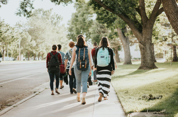 A group of backpacked people walks along a sunny tree-lined sidewalk beside a quiet street.