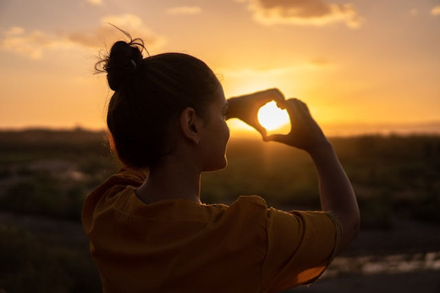 Silhouette of a woman at sunset framing the sun with her hands in a heart shape, warm golden sky and calm mood.