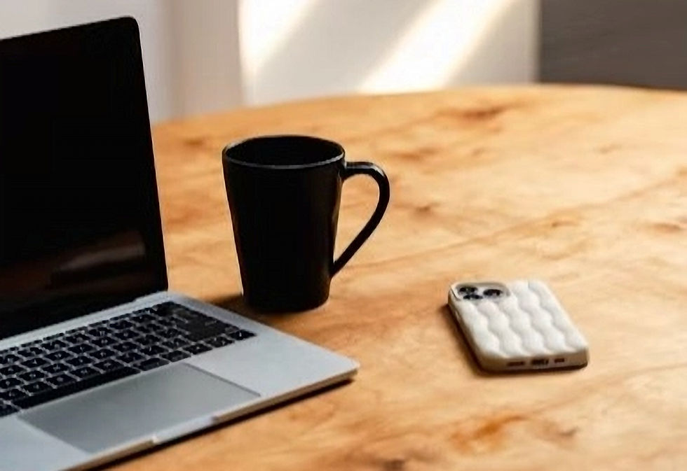 A laptop, a black coffee mug, and a smartphone with a white case rest on a wooden table bathed in natural light.