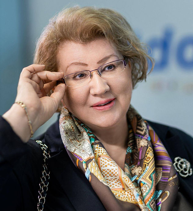A woman adjusts her glasses, smiling in an office setting. She wears a patterned scarf and dark blazer with a brooch. Background is blurred.