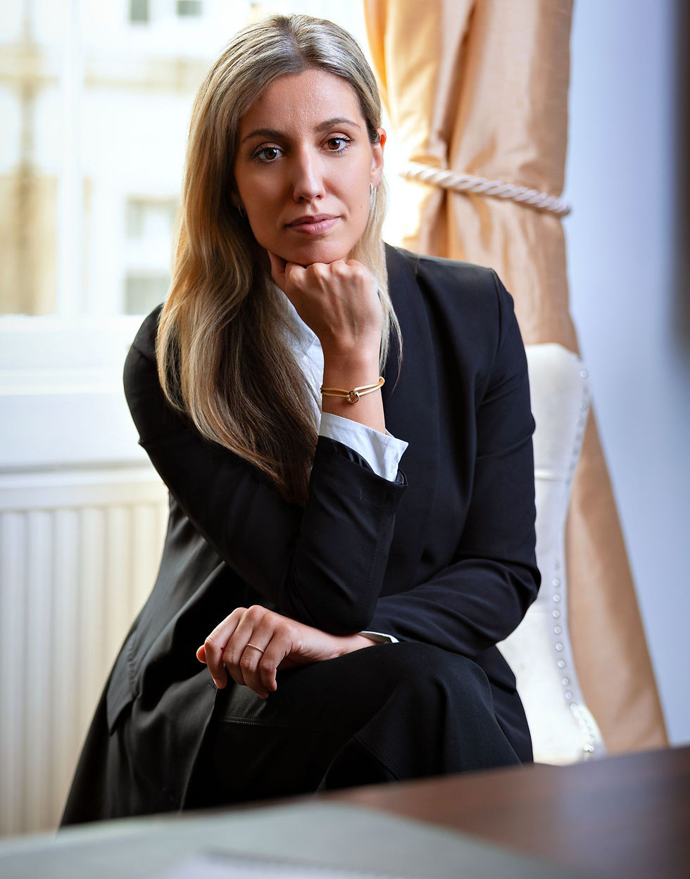 A woman in a black suit sits pensively in an elegant room, resting her chin on her hand. Beige curtains and a large window in the background.