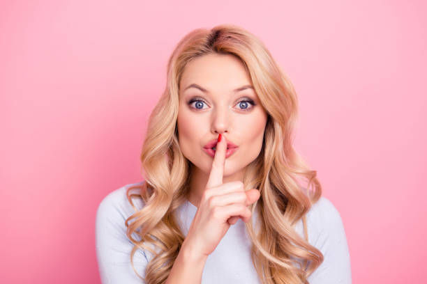 A blonde woman with wide eyes holds her finger to her lips in a "shh" gesture, standing against a pink background.
