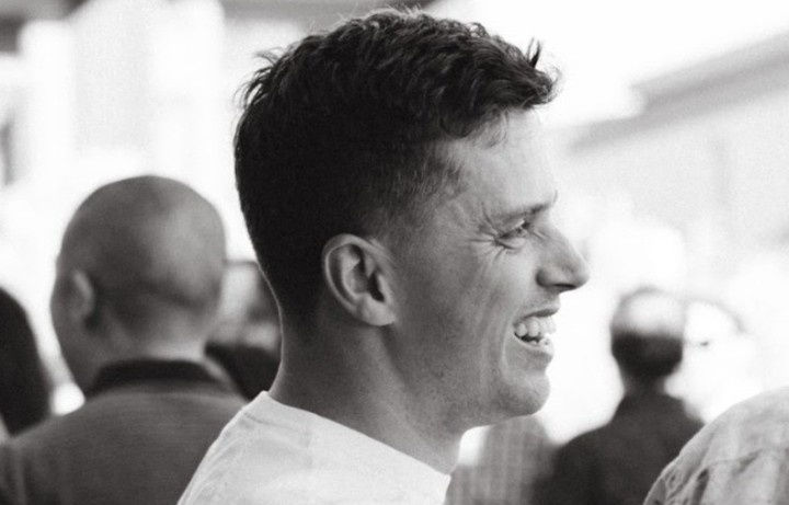 Man smiling in profile, wearing a white shirt, in a grayscale image with blurred people in the background, creating a joyful mood.