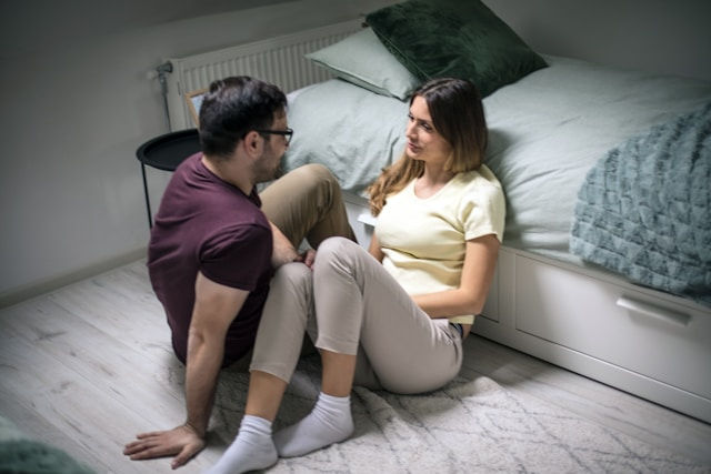 Man and woman sit on the bedroom floor beside a bed, talking closely in a calm, intimate moment.