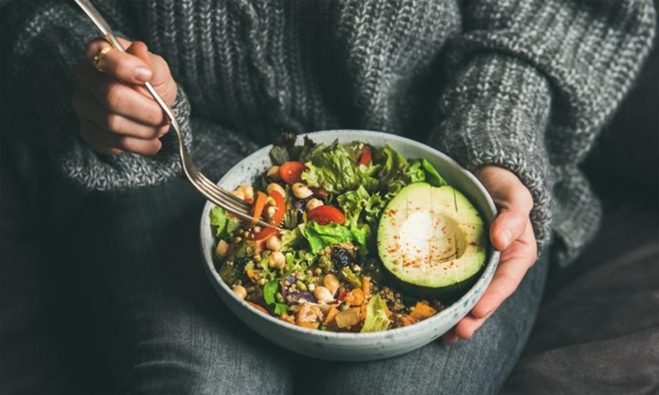Person in gray sweater holds a bowl of salad with avocado, cherry tomatoes, and greens. Fork in hand, cozy setting, warm tones.