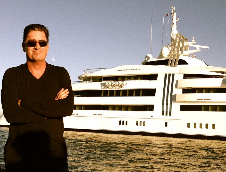 Man in sunglasses stands with arms crossed in front of a large white yacht named Vibrant Curiosity, on a sunny day at sea.