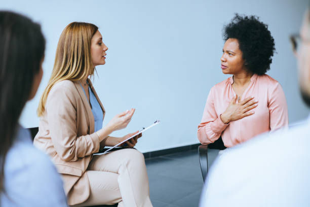 Two women engaged in a conversation, one holding a clipboard, the other with a hand on her chest. Neutral-toned office setting.