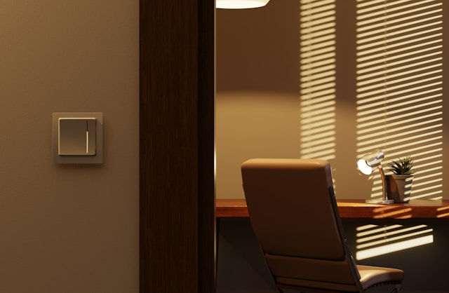 Modern office corner with brown chair, wooden desk, lamp and plant, striped window-blind shadows, and a wall light switch.