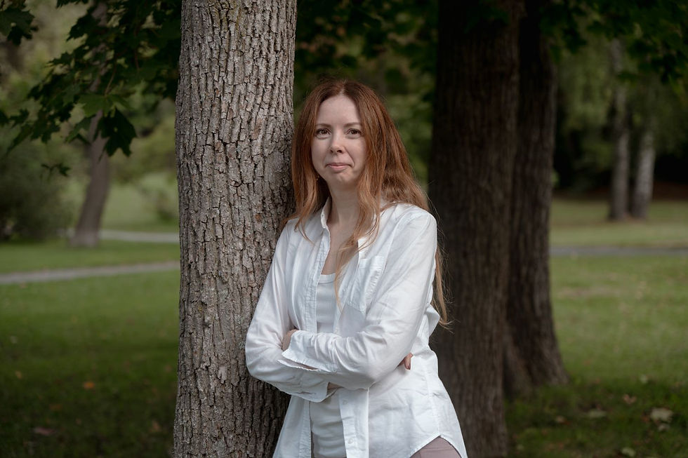 Woman in a white shirt stands with arms crossed against a tree in a park setting, surrounded by greenery, expressing a calm demeanor.