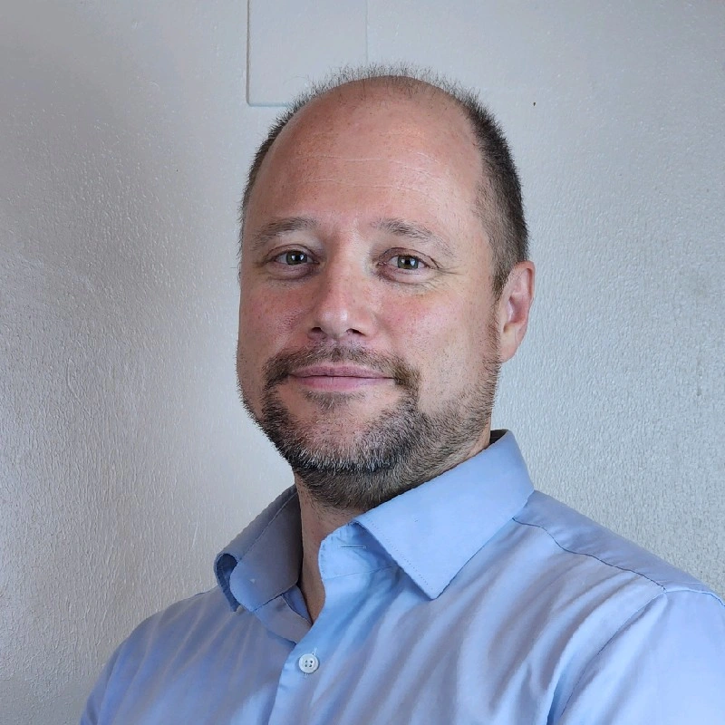 Man with a beard wearing a navy suit and shirt, smiling slightly. Neutral gray background, conveying a calm and professional mood.