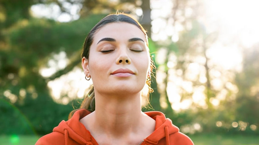 Woman in a red hoodie meditates with closed eyes, surrounded by sunlit trees. She appears calm and relaxed.