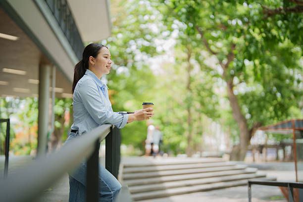 Woman in a light blue shirt holds a coffee cup, leaning on a railing outdoors by steps in a leafy park.