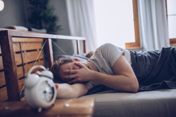 Man lying in bed covering face, appearing tired. White alarm clock on wooden nightstand. Soft natural light from window, cozy setting.