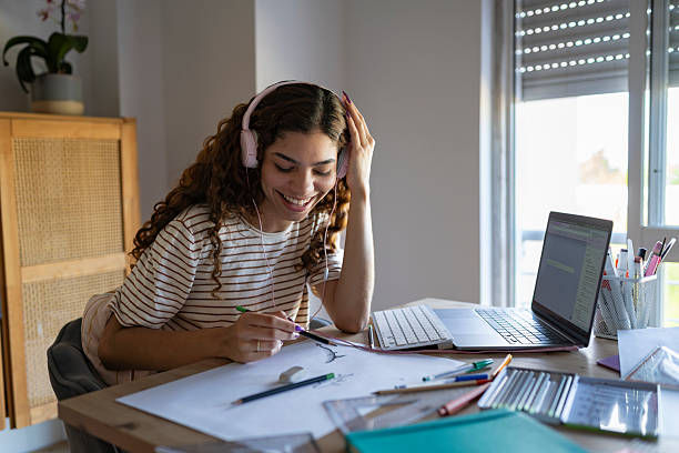 Smiling woman in headphones studies at a desk with laptop, notebook and pens by a sunlit window at home
