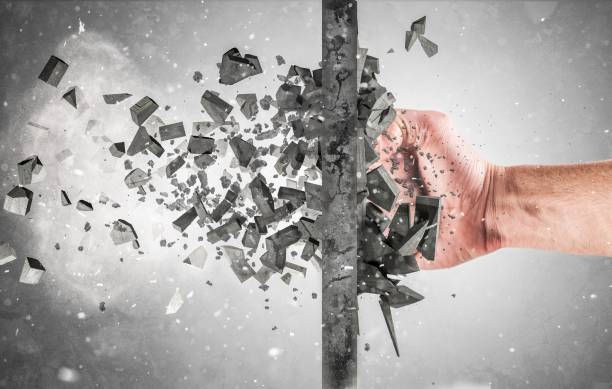 Fist punching through a dark wall, shattering it into flying shards against a gray background, symbolizing force and breakthrough