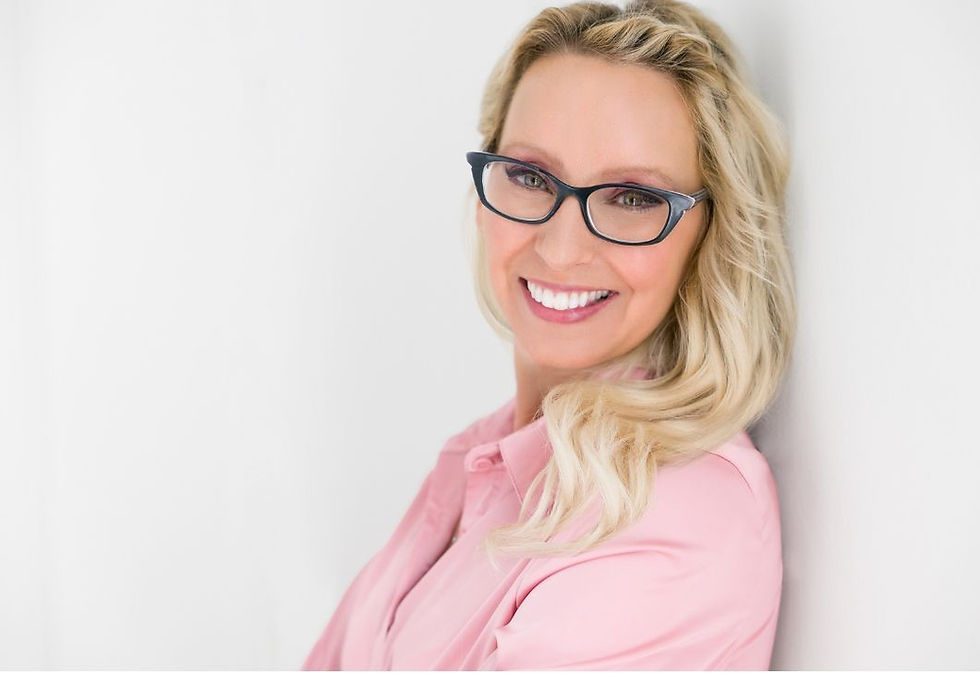 Smiling woman with glasses and blonde hair in a pink shirt stands against a white wall, exuding a cheerful mood.