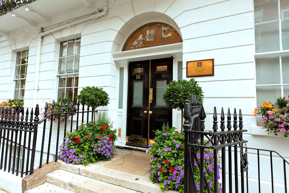 White townhouse with black door marked "10". Surrounded by colorful flowers and greenery, wrought iron fence, and a golden plaque.