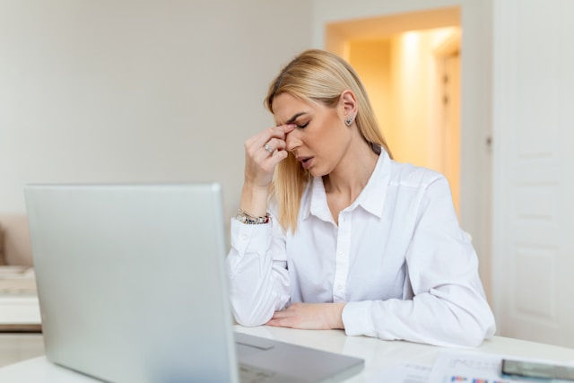 Blonde woman in a white shirt sits at a laptop, rubbing her forehead in a bright home office, looking stressed and tired.