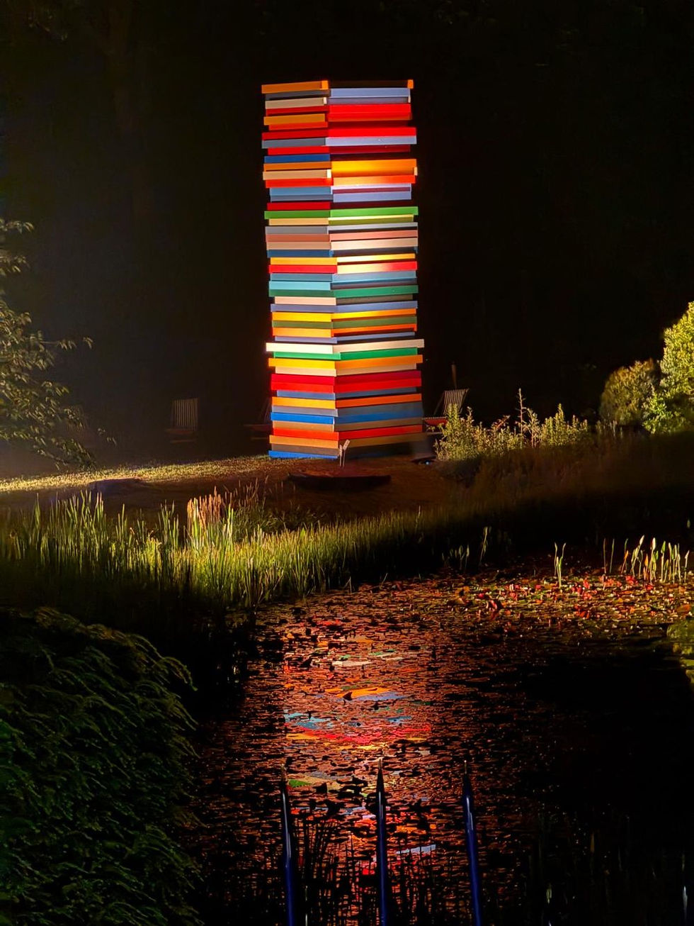 Colorful stacked lighted tower at night beside a pond, its bright stripes reflecting in the water.