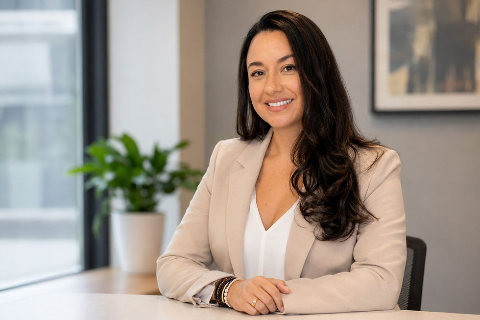 Smiling woman in a beige blazer sits at a desk in a bright office with a window and potted plant in the background.